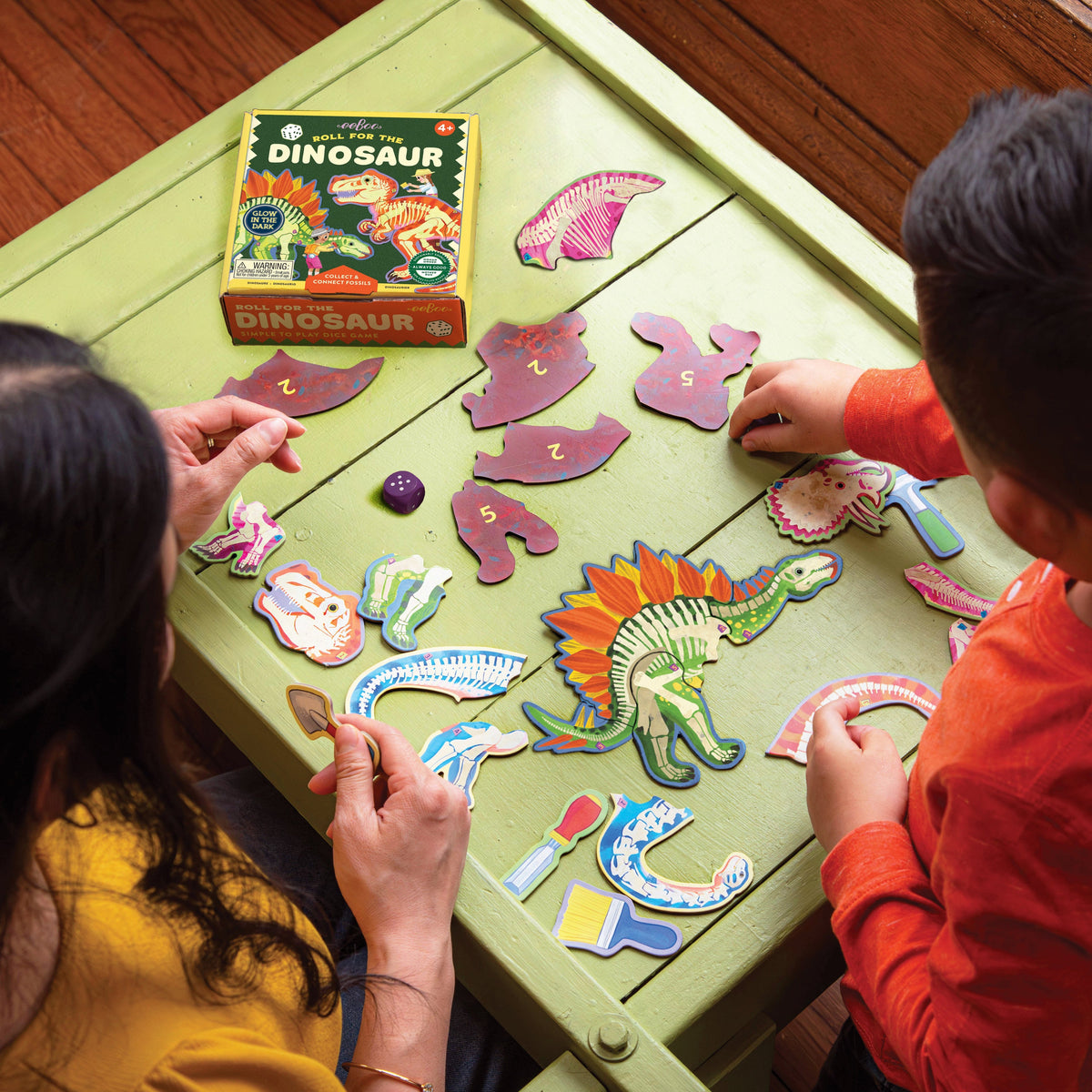 A child and an adult sitting at a table playing the Dinosaur Dice Game by Eeboo, assembling colorful dinosaur fossil puzzle pieces around the game box.