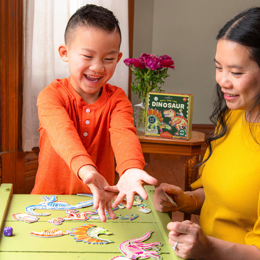 A child and an adult playing the Dinosaur Dice Game by Eeboo together at a table, with colorful dinosaur fossil puzzle pieces spread out and the game box visible in the background.