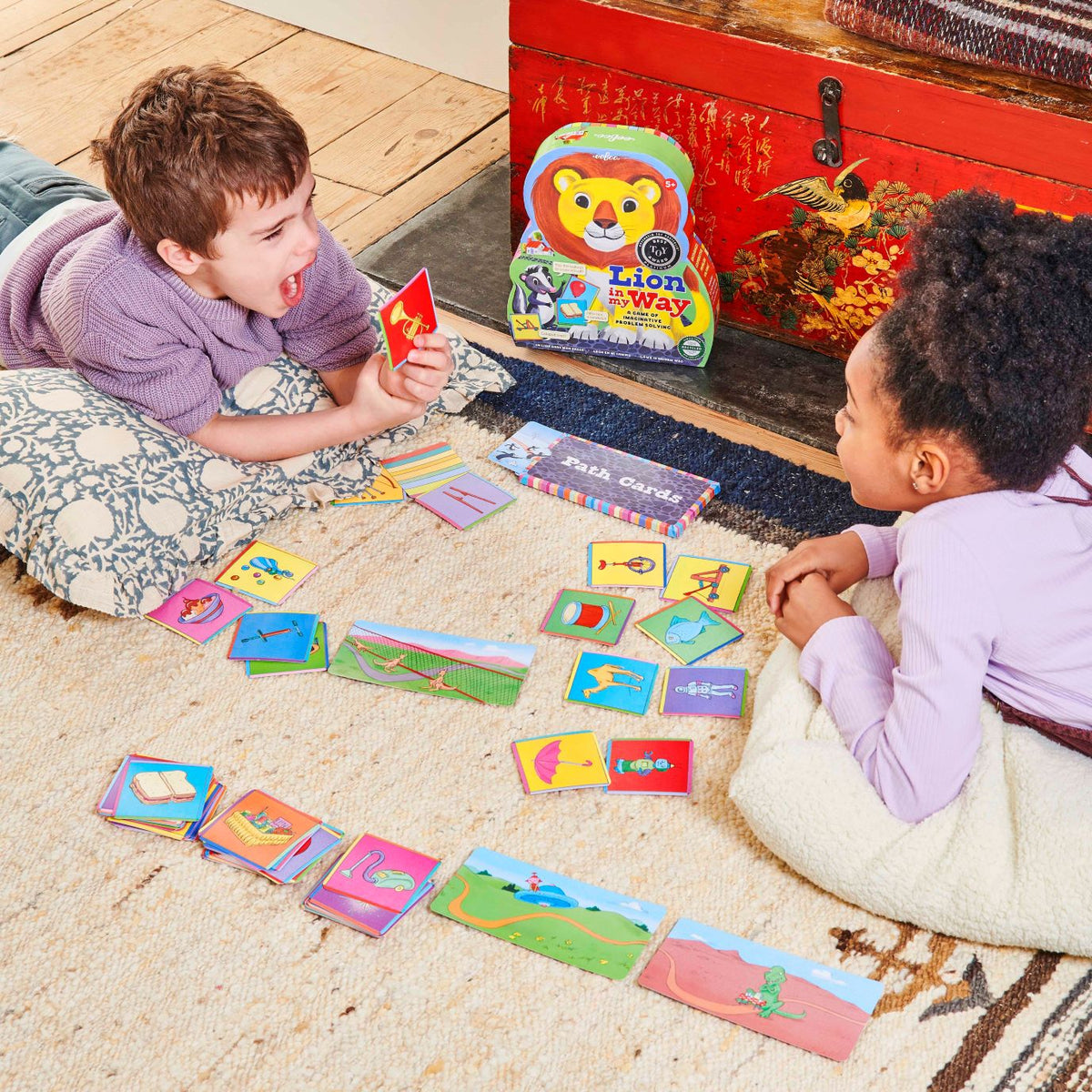 Two children playing with colorful cards on the floor next to a red toy chest.