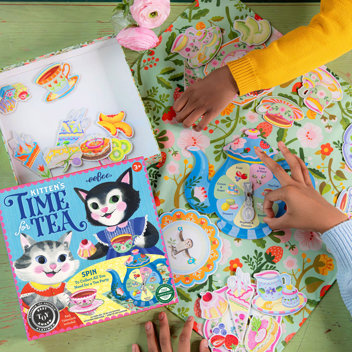 Kitten's Time for Tea Spinner Game by eeBoo box and game components laid out on a floral tablecloth, with a child spinning the teapot spinner and colorful cutouts of tea party treats nearby.