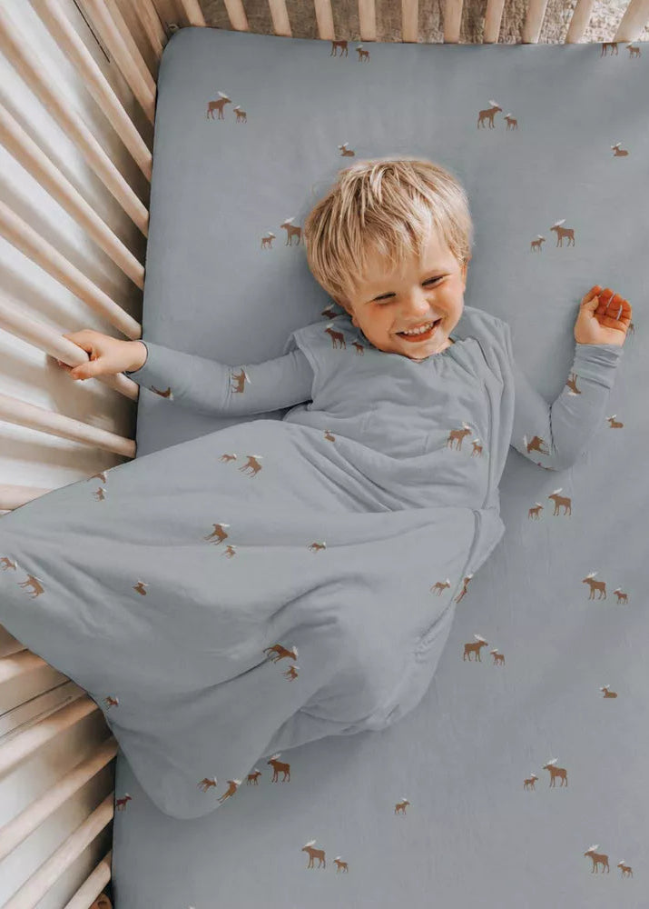 A smiling toddler lies on their back in a wooden crib, wearing a long-sleeve sleep sack and matching fitted crib sheet. The fabric is a dusty slate blue with a small brown and white deer print, set against a neutral cream-colored crib.