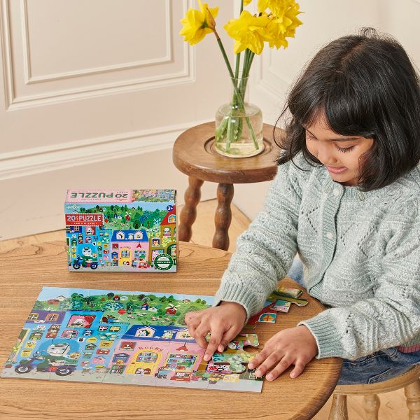 A child sitting at a wooden table working on the 20 Piece Puzzle - Yumi's Village by Eeboo, with the colorful puzzle box resting on the table above the completed village scene.