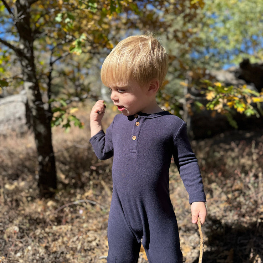 A toddler with blonde hair wearing a dark navy long-sleeve footless onesie with a three-button henley collar, standing outdoors in a wooded area with autumn trees.