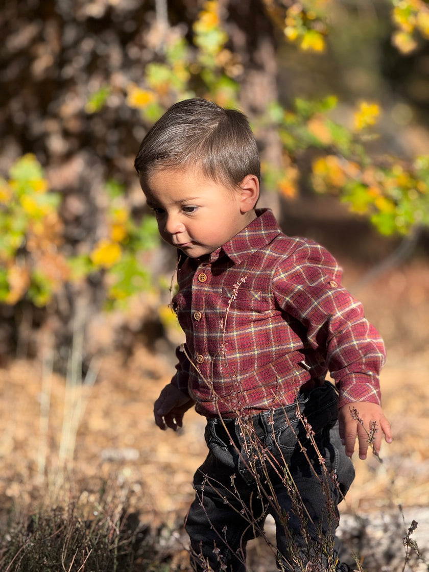A young boy wearing a red and green plaid button-down shirt and dark pants, standing outdoors in a natural setting with dry brush and blurred trees.