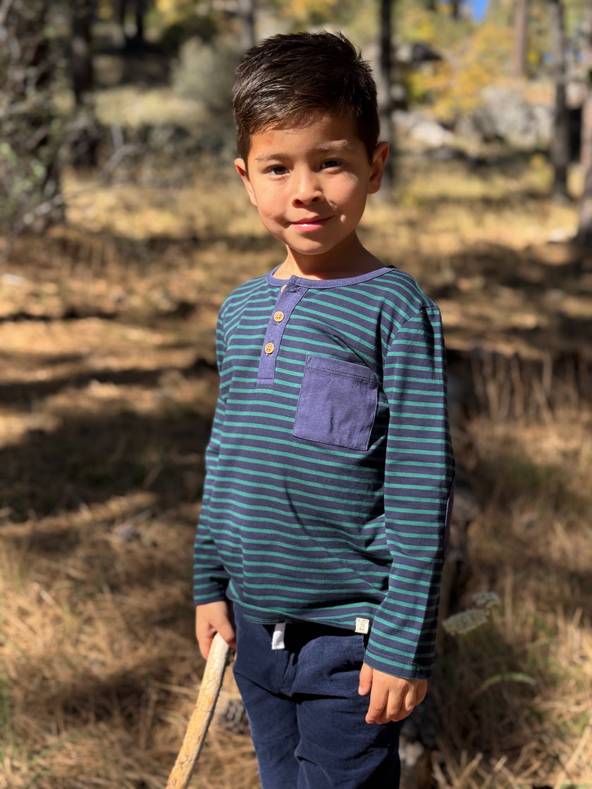 A young boy wearing a navy and green striped long-sleeve henley shirt with a purple pocket, standing outdoors in a wooded setting.