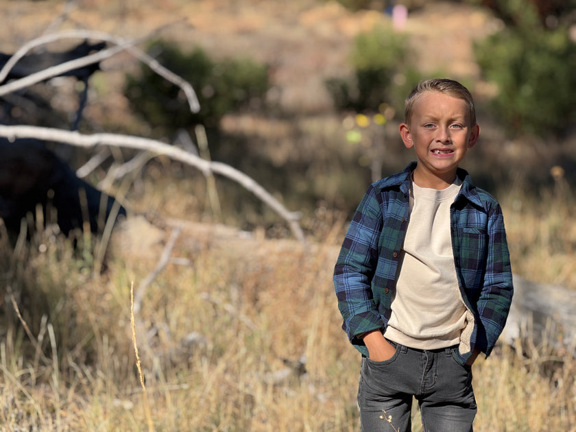 A young boy standing outdoors in a dry grassy field, wearing a blue and green plaid flannel shirt over a cream t-shirt and dark gray jeans.