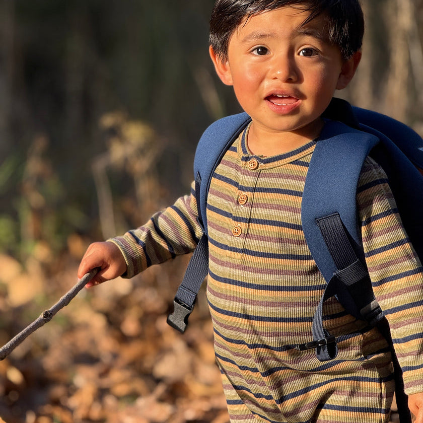 A toddler boy wearing a blue backpack and a beige and brown striped long-sleeve henley jumpsuit.