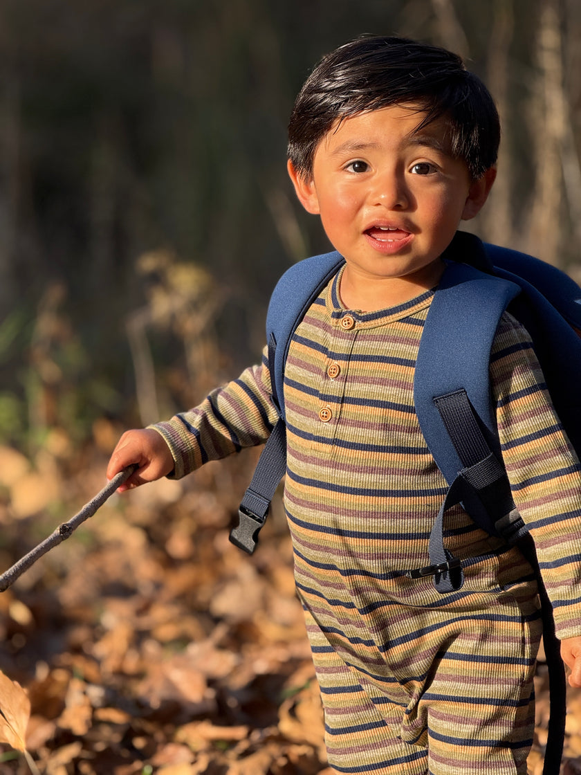 A toddler boy wearing a blue backpack and a beige and brown striped long-sleeve henley jumpsuit.