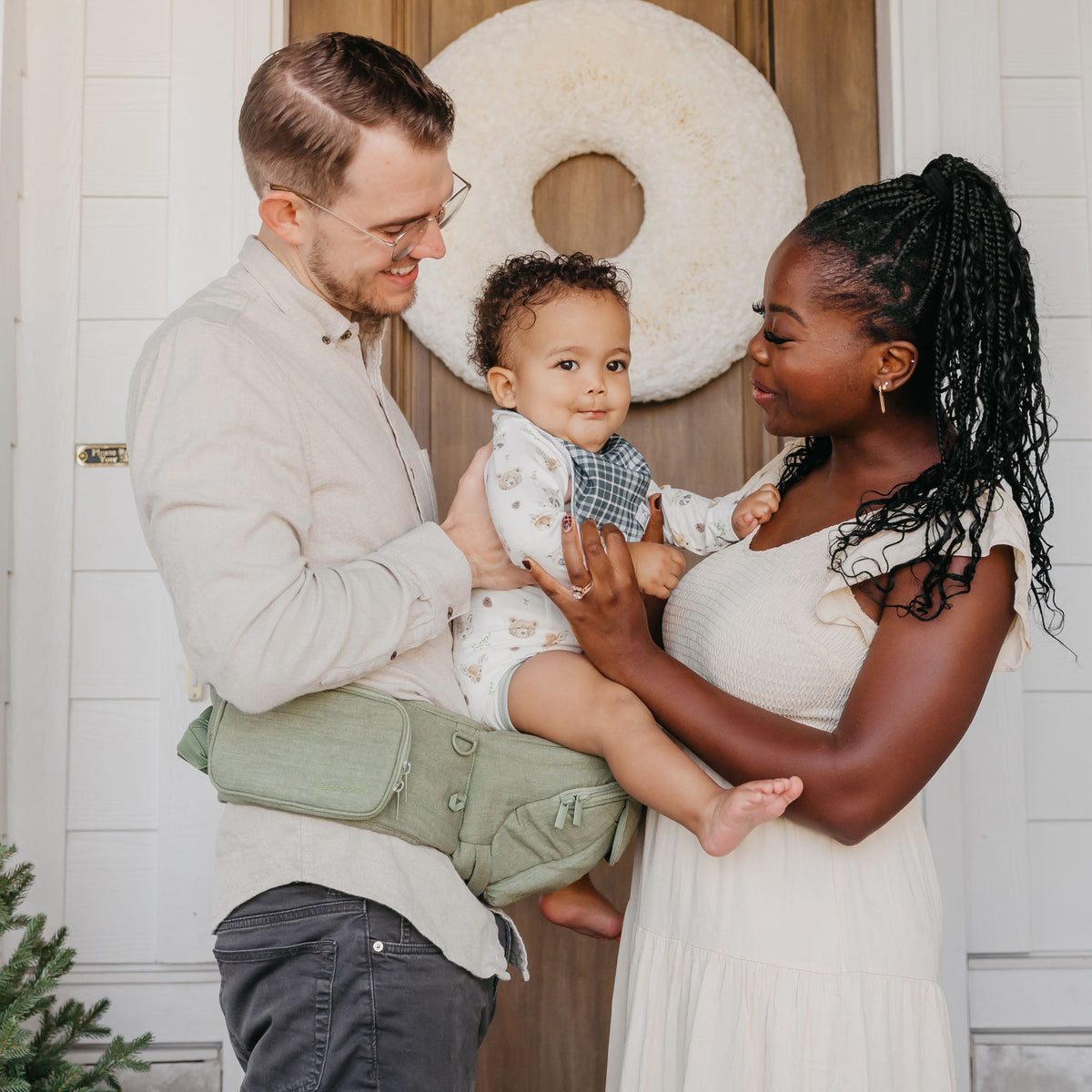 Family of three standing in a doorway with a decorative wreath, one person holding a baby.