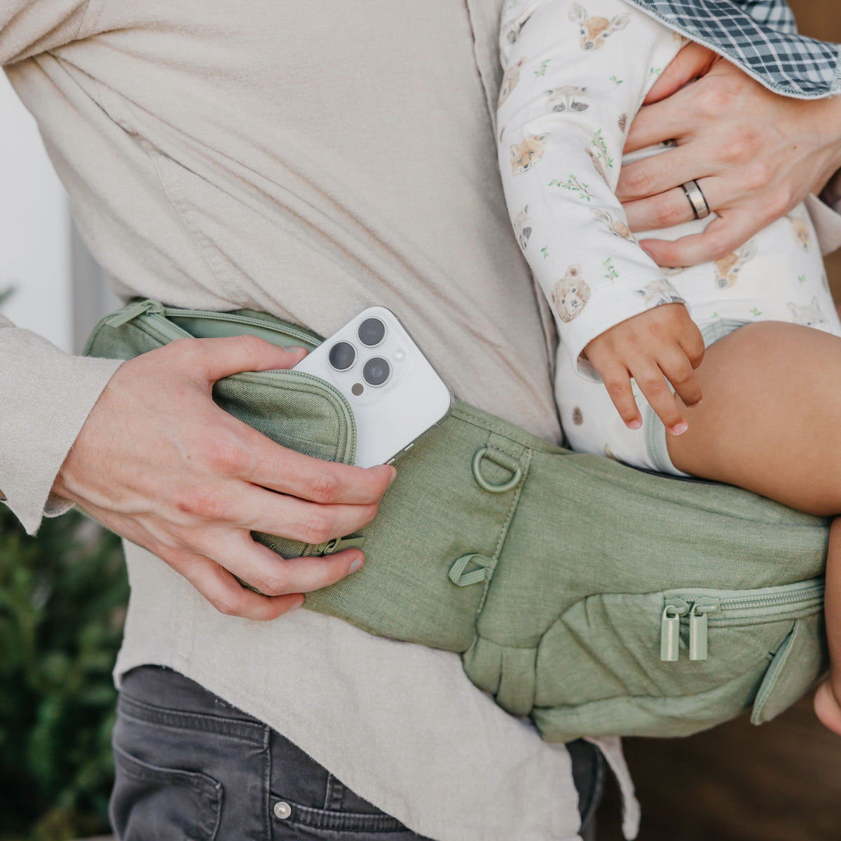 Person holding a phone with a green baby carrier and a child in a floral outfit.
