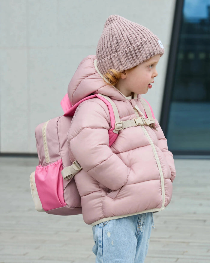 A child wearing a light pink puffer jacket, a matching pink knit beanie, and a pink backpack, standing outdoors against a gray wall.
