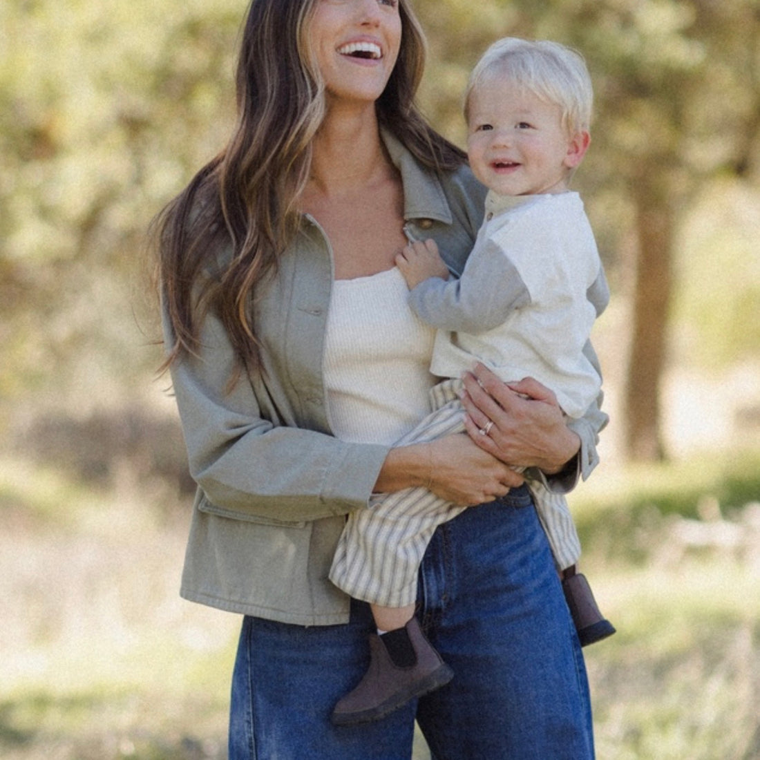 A woman outdoors holding a smiling toddler. The woman wears an olive green utility jacket over a white top and blue jeans, while the child wears a white long-sleeve shirt and light striped pants.
