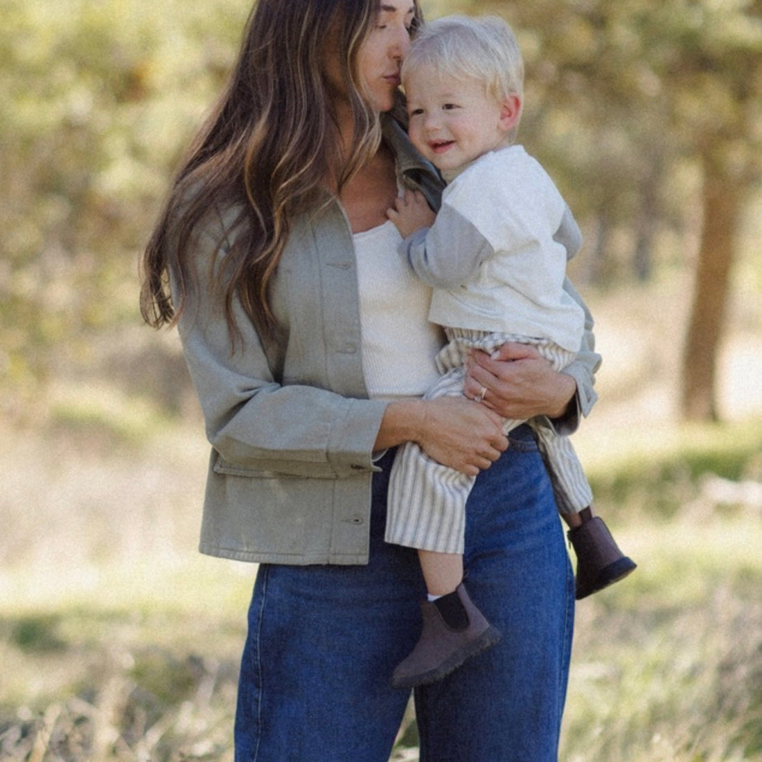 A woman outdoors holding a baby. The woman is wearing a light sage green button-up shirt over a white top and blue jeans.