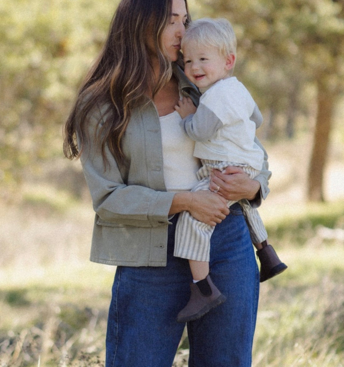 A woman outdoors holding a baby. The woman is wearing a light sage green button-up shirt over a white top and blue jeans.