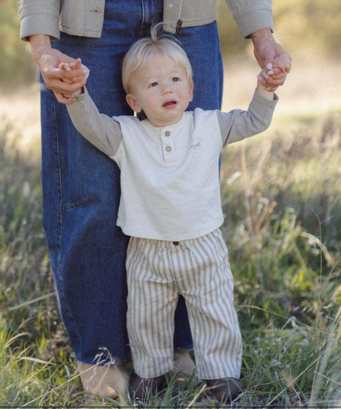 A toddler standing outdoors in a grassy field holding hands with an adult wearing blue jeans and a beige jacket. The child is wearing a light beige henley shirt with gray sleeves and beige-and-white vertically striped pants.