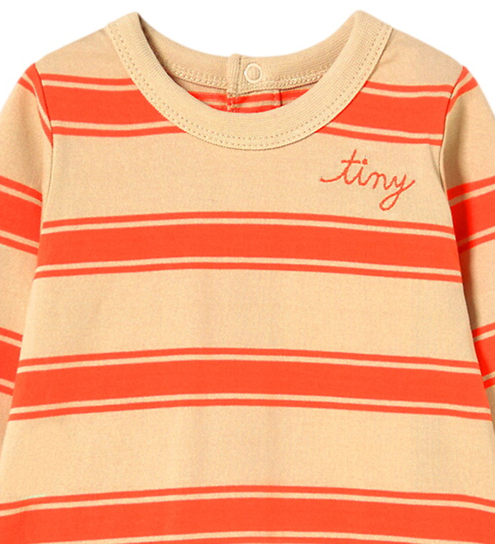 A close-up of a baby long-sleeve top with horizontal orange and white stripes on a beige background, featuring the word 'tiny' embroidered in orange script on the chest.
