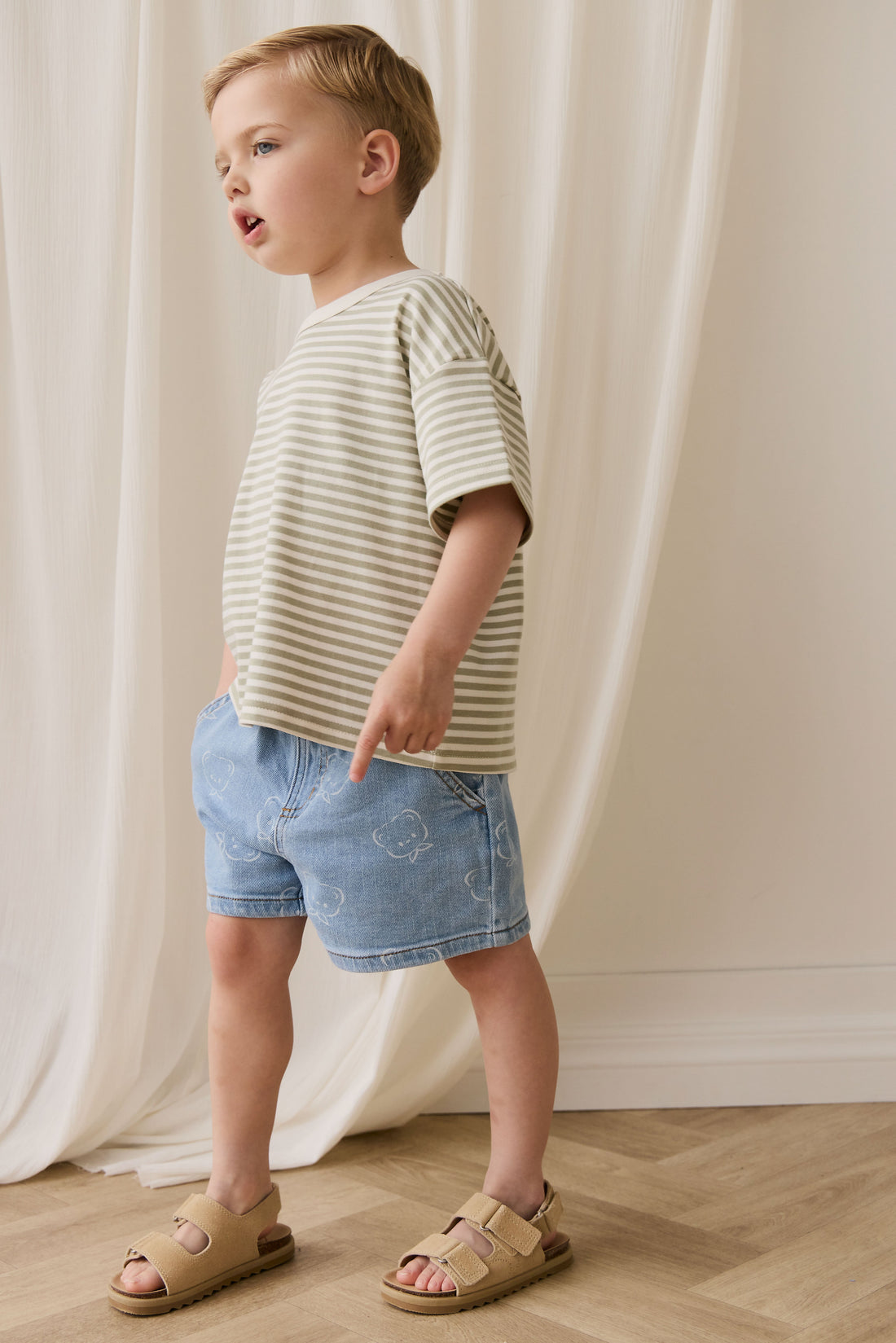 Child wearing a striped shirt and denim shorts standing against a white curtain.