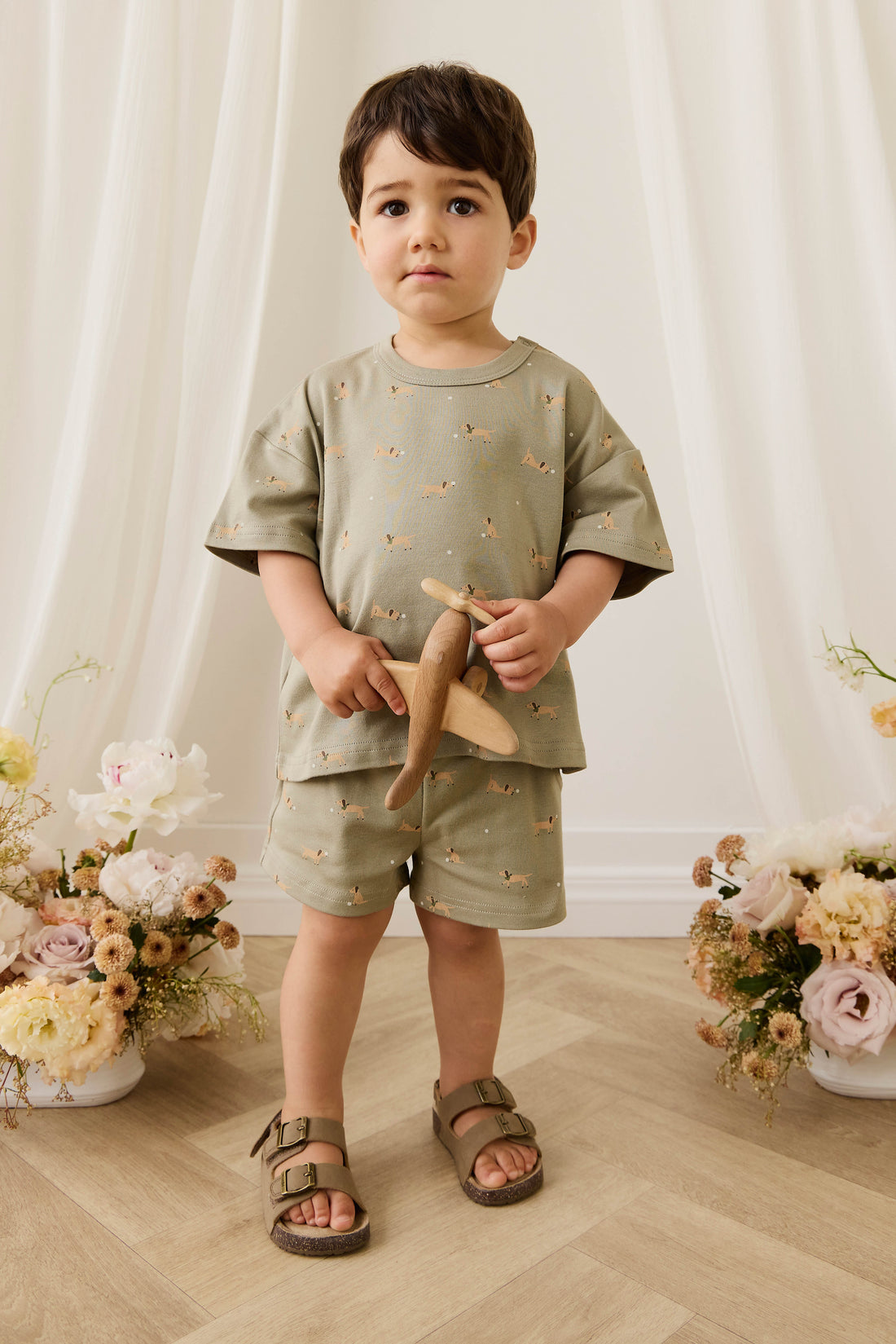 Child wearing a green outfit with floral patterns, standing in front of white curtains with floral decorations on either side.