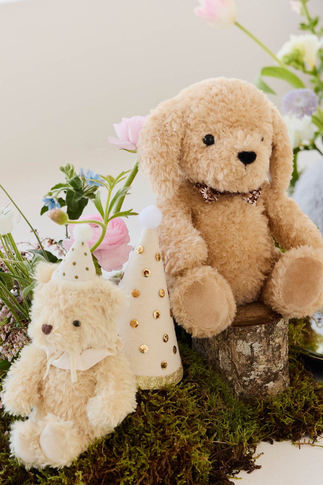 An eye-level studio product shot of the "Snuggle Bunnies - Honey the Puppy by Jamie Kay" plush toy sitting upright on a small wooden log stump. The puppy is made of soft, textured honey-toned faux fur with long floppy ears, shiny dark brown button eyes, a stitched dark brown nose, and a checkered ribbon collar. The rustic setting is styled with patches of green moss at the base and a party hat with gold foil polka dots positioned beside the toy.