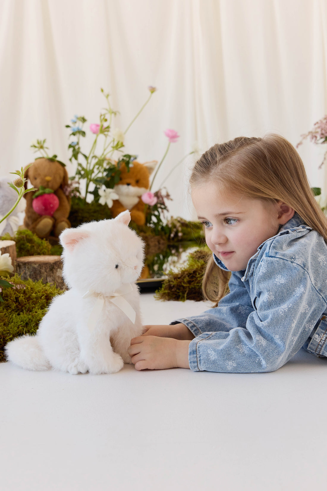 A young child with long blonde hair, wearing a denim jacket, smiles while holding a fluffy white plush kitty toy against a neutral background.