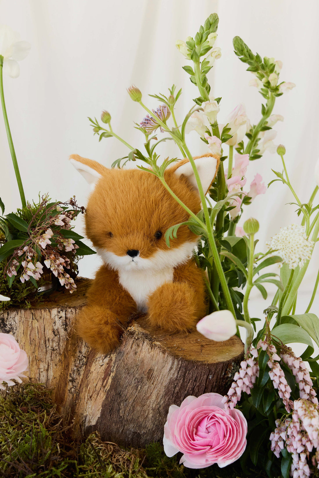 A plush toy fox sitting on a rustic wooden stump surrounded by blooming spring flowers and lush greenery.