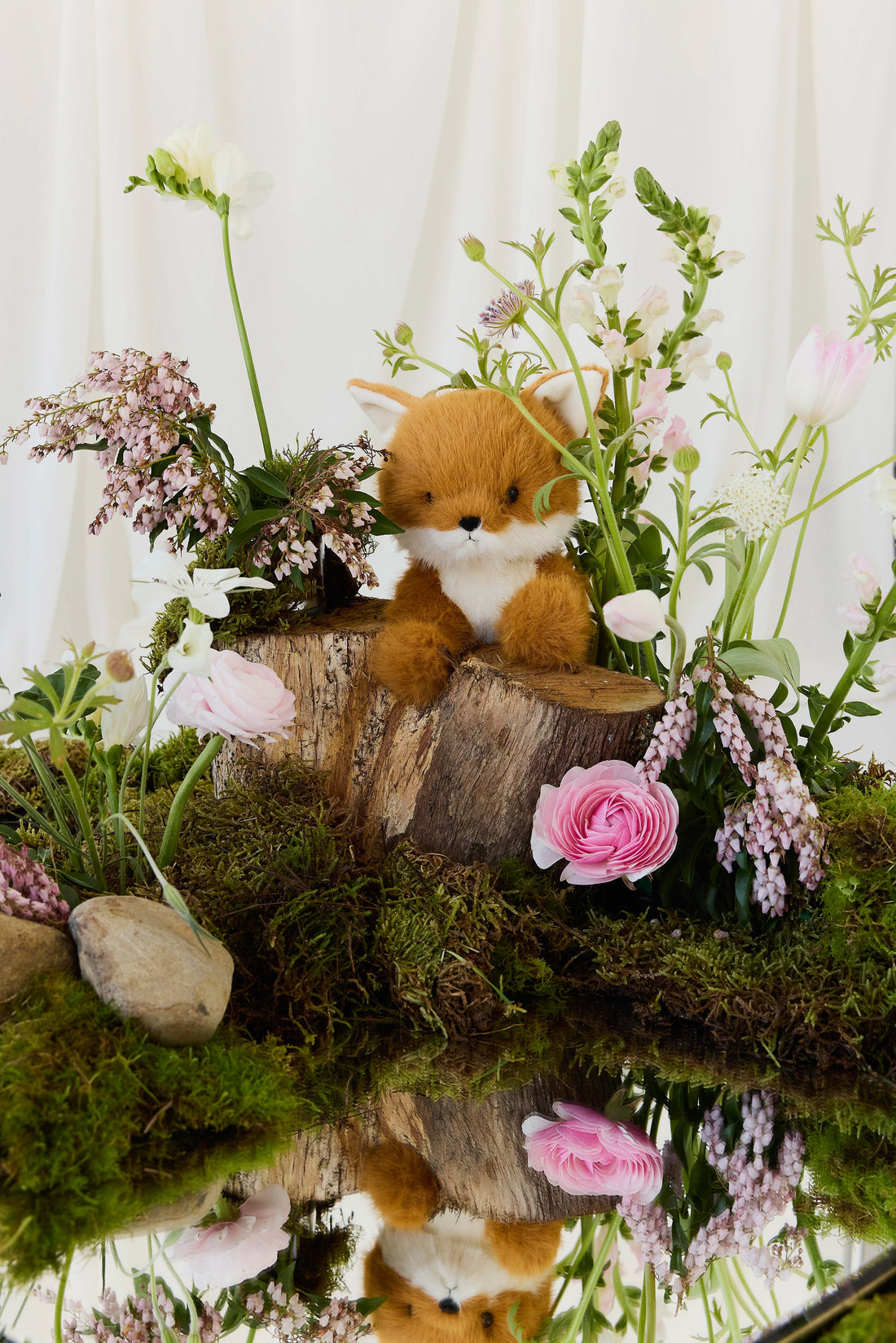 A plush toy fox sitting on a rustic wooden stump surrounded by blooming spring flowers, greenery, and a reflective surface below.