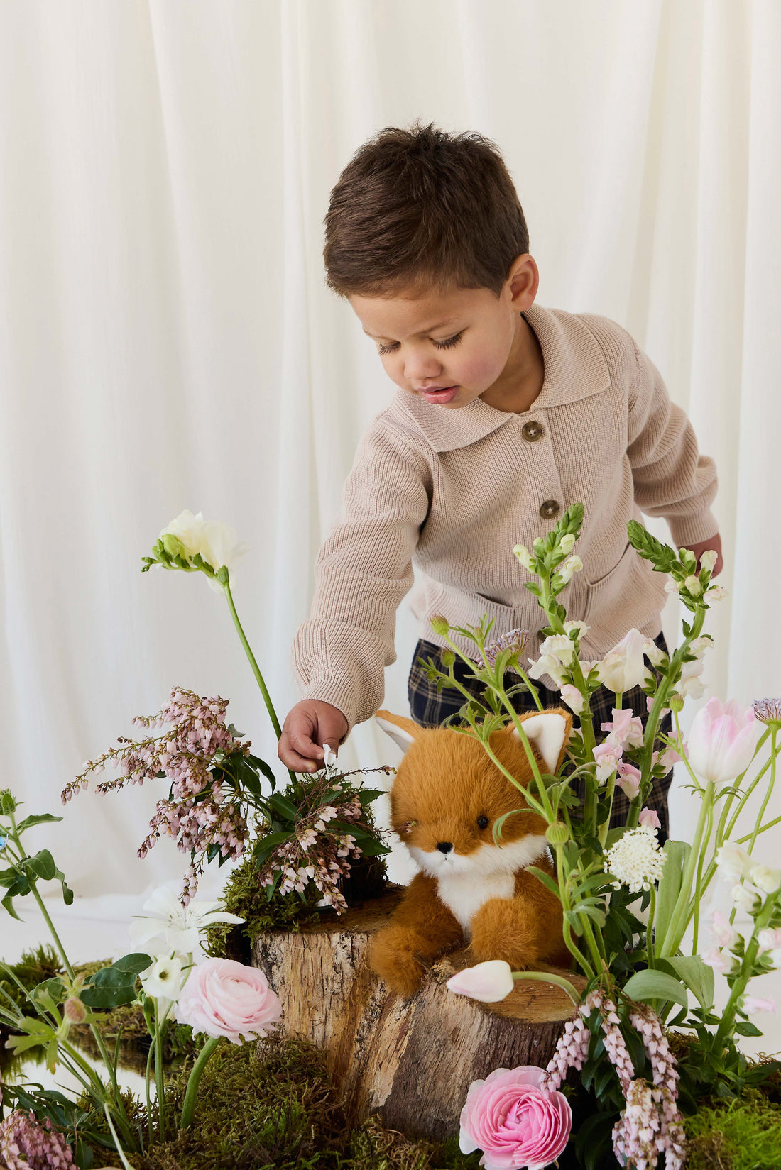 A young child wearing a beige knit cardigan reaches gently toward a plush fox toy nestled among blooming spring flowers and greenery on a rustic wooden stump.