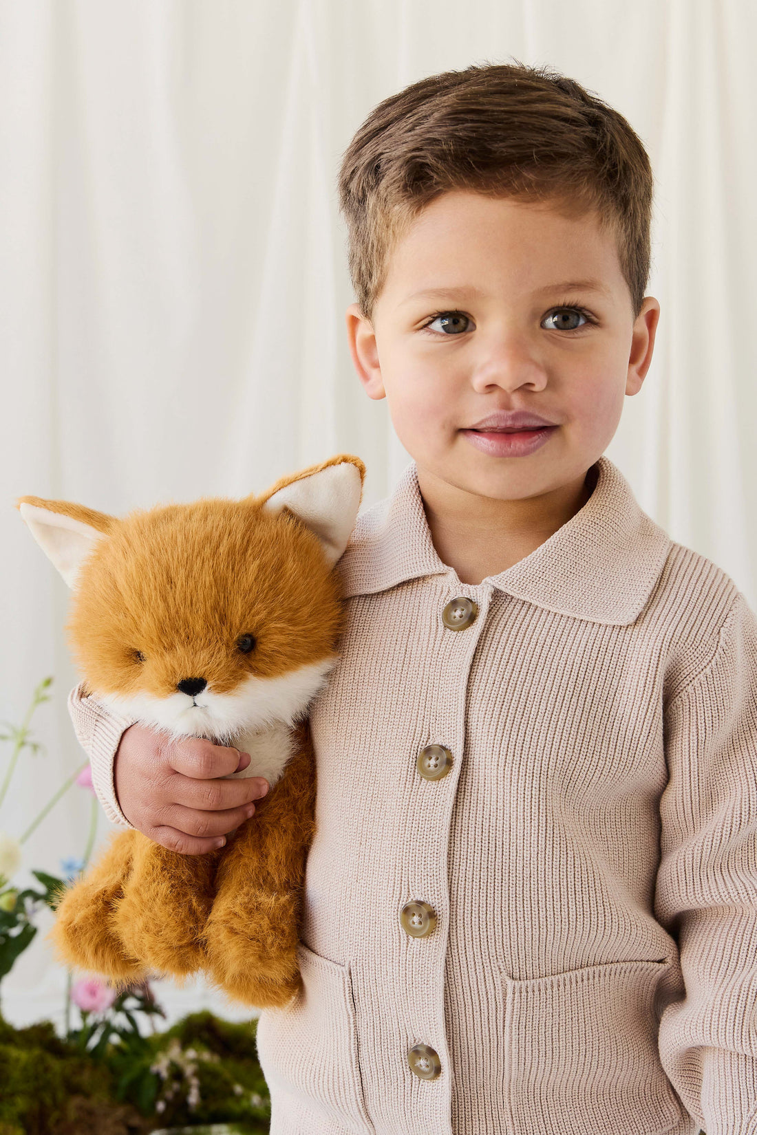 A young child smiling gently while wearing a beige knit button-down cardigan, holding a plush fox toy against a soft, neutral-toned background.