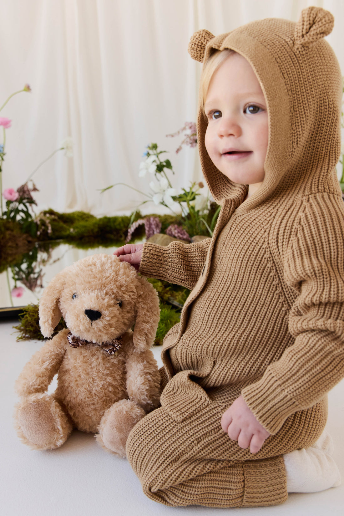An eye-level studio portrait of a toddler sitting indoors against a light-colored backdrop, wearing a matching textured brown knit romper with an attached hood featuring small, upright bear ears and a front kangaroo pocket. Beside the toddler, the plush honey-toned puppy toy with floppy ears and a checkered ribbon collar sits upright against the backdrop. The soft, natural lighting illuminates the scene, which is styled with delicate floral arrangements and moss at the base.
