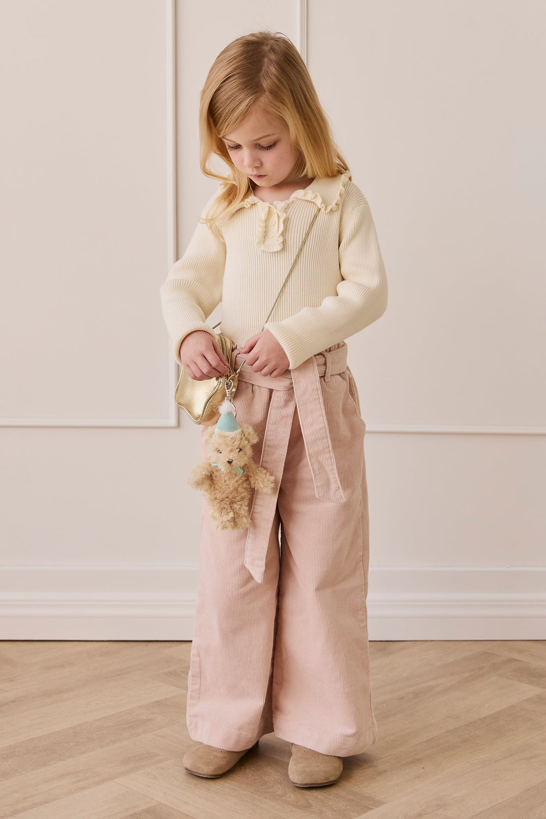 A young girl with blonde hair and a cream-colored knit top holding a gold star-shaped bag and the fluffy beige teddy bear keyring wearing a light blue party hat and matching ribbon.
