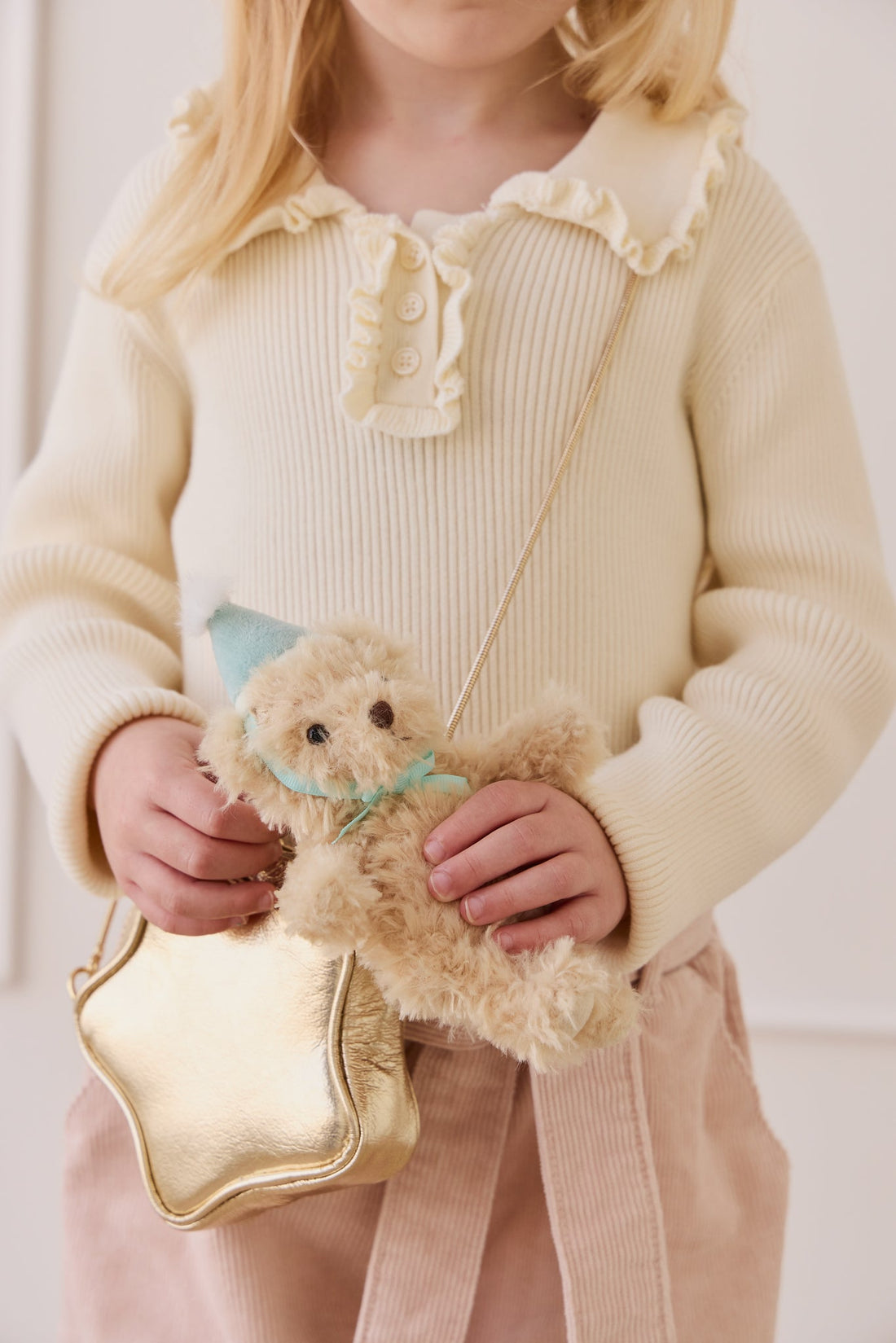 A young girl with blonde hair and a cream-colored knit top holding a gold star-shaped bag and the fluffy beige teddy bear keyring wearing a light blue party hat and matching ribbon.