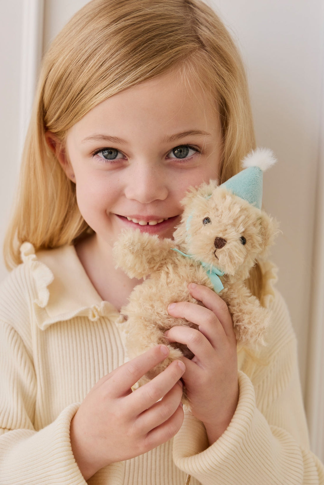 A young girl with blonde hair and a cream-colored knit top smiling warmly as she holds a fluffy beige teddy bear keyring wearing a light blue party hat and a matching ribbon.