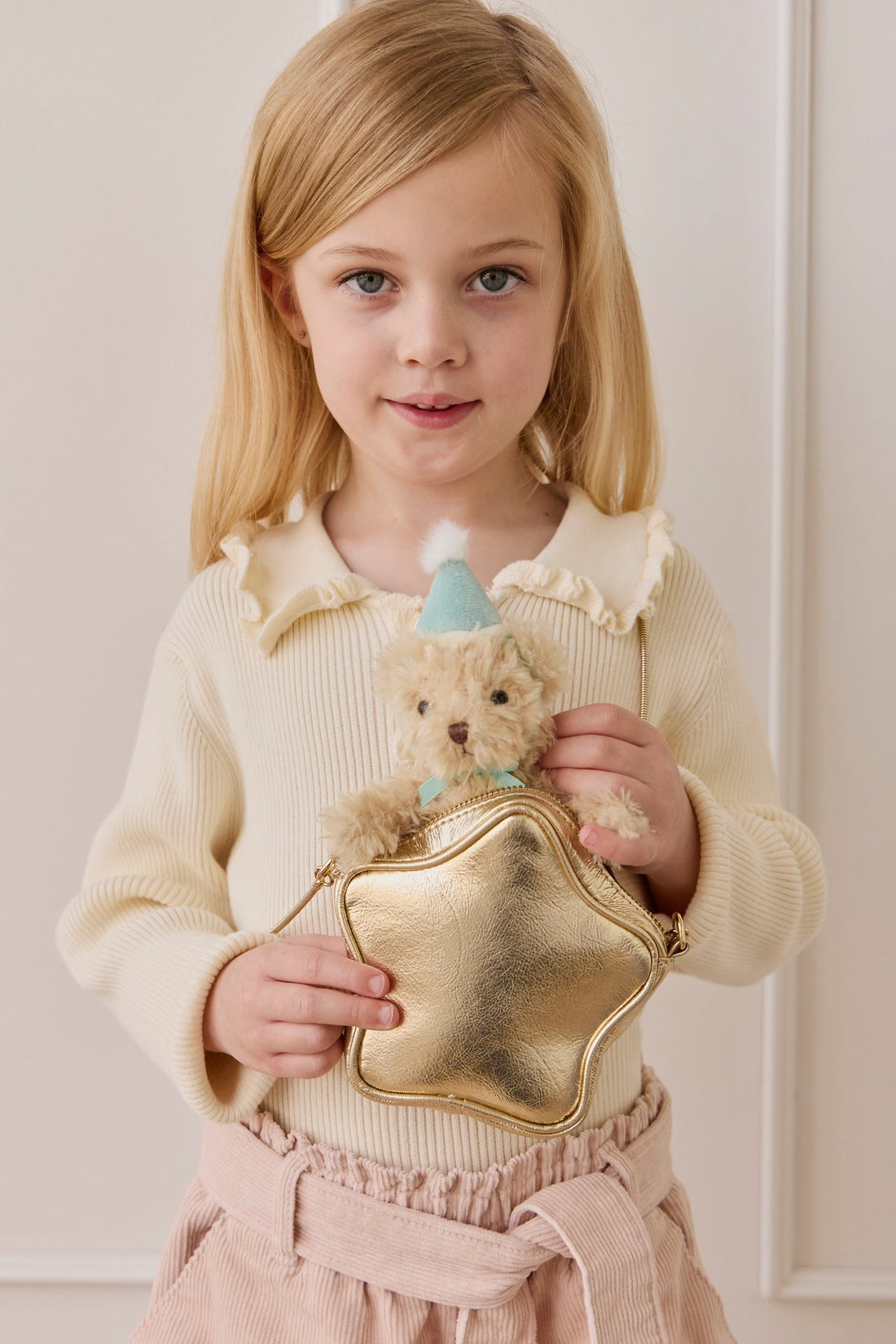 A young girl with blonde hair and a cream-colored knit top holding a gold star-shaped bag and the fluffy beige teddy bear keyring wearing a light blue party hat and matching ribbon.