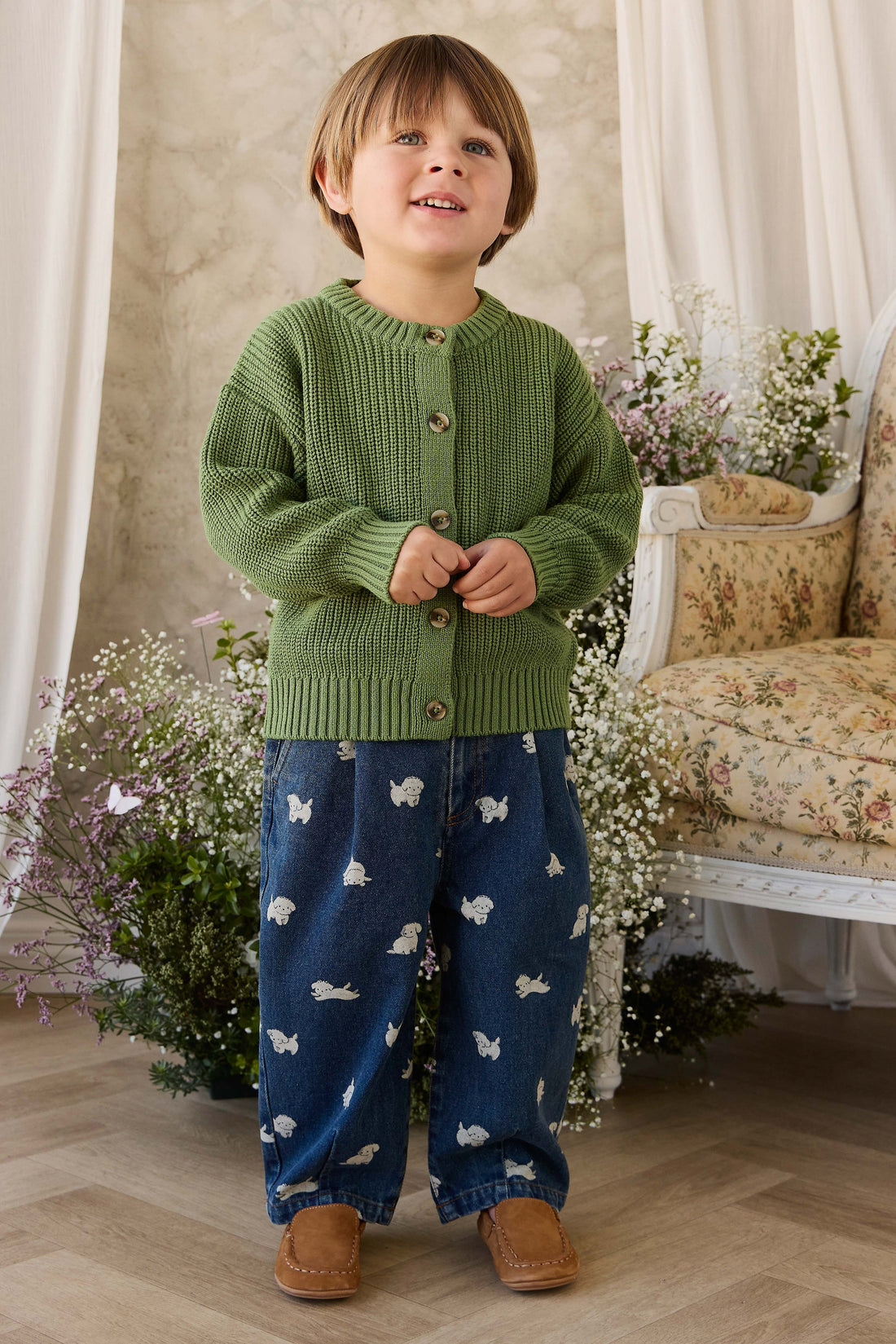 Child wearing a green knitted cardigan and blue pants with animal patterns, standing in a decorated room.