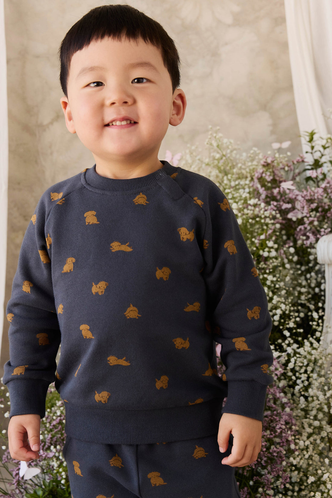 A young boy wearing a navy blue sweatshirt and matching pants set, both decorated with small mustard-gold dog silhouettes, standing in front of a soft-focus floral background.