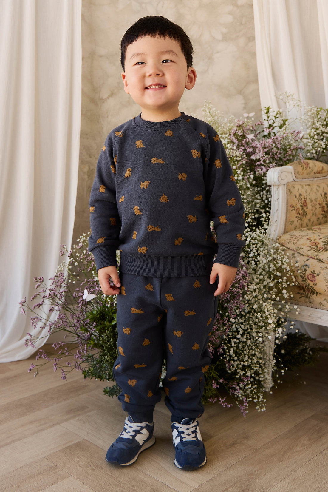 Child wearing a navy sweater and pants with small patterns, standing in a decorated room with flowers and a chair.