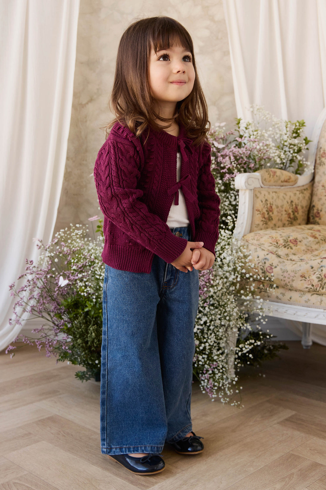 Young girl wearing a maroon cardigan and blue jeans standing in a decorated room with flowers and a couch.