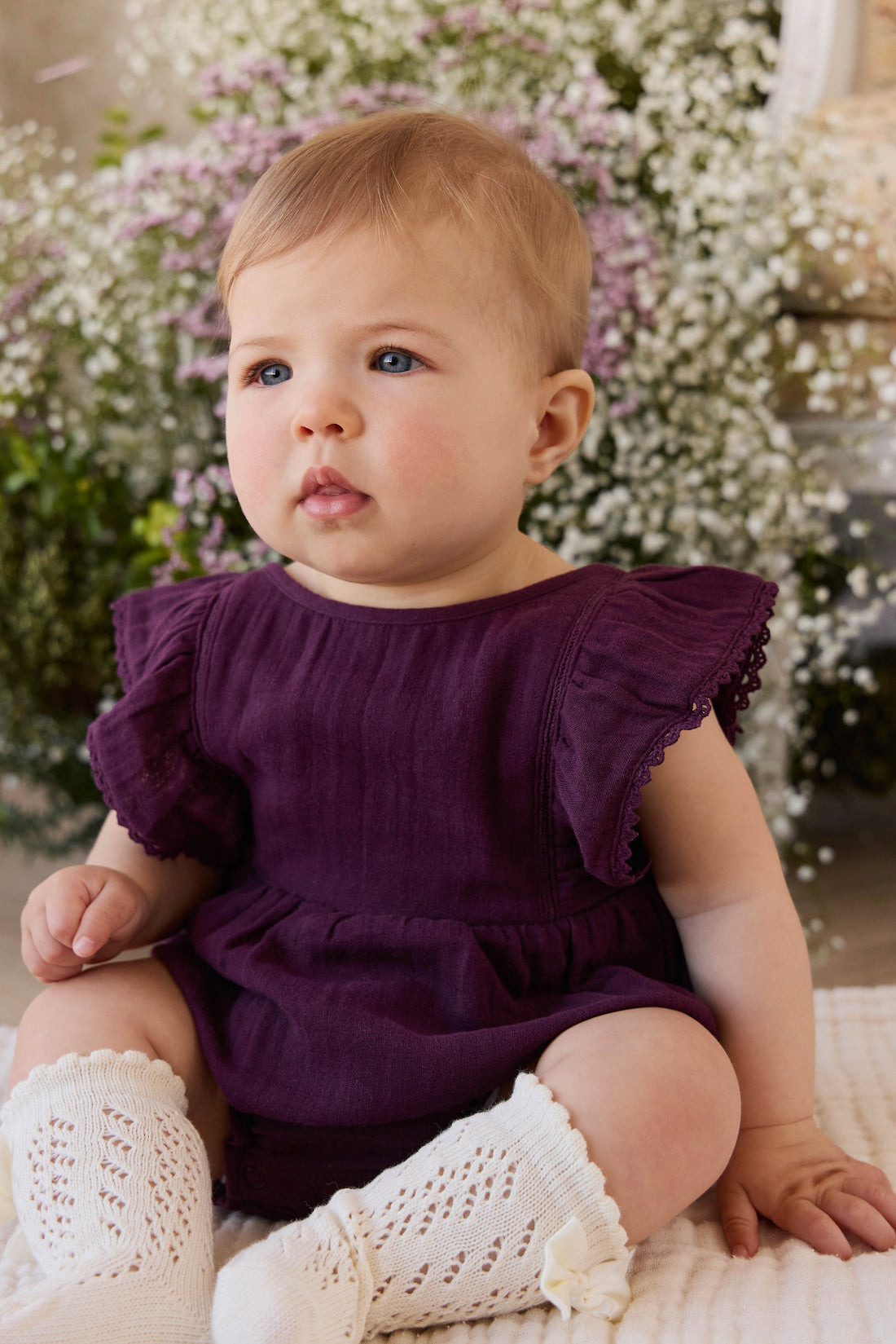 Baby in a purple dress with ruffled sleeves sitting on a floral background