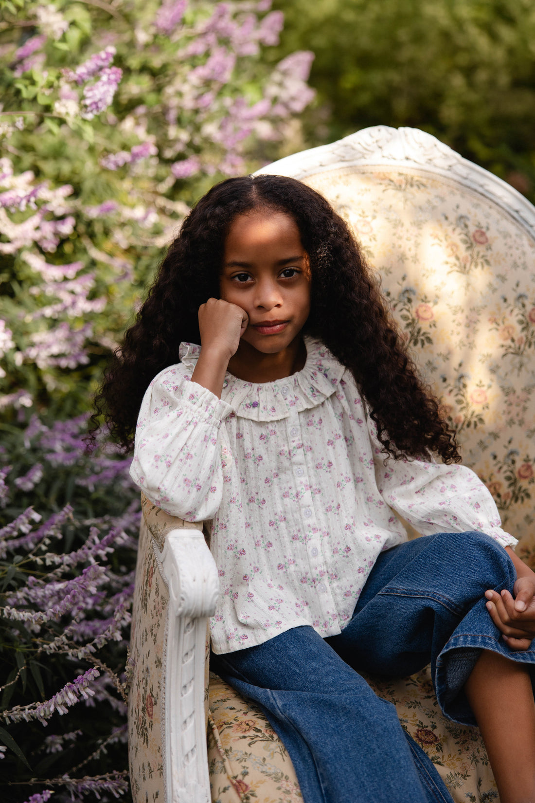 A young girl with long curly dark hair sits in a floral armchair outdoors, wearing a white long-sleeve blouse with a small pink floral print and blue jeans. Purple flowers and green foliage are visible in the background.