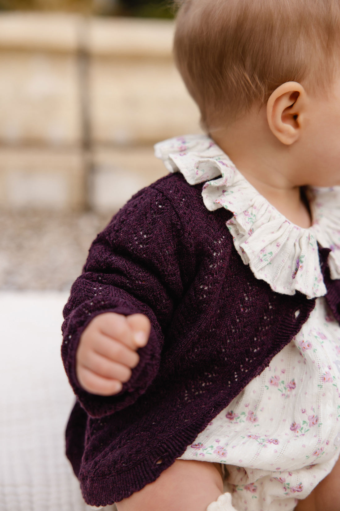 Baby wearing a dark purple cardigan over a floral outfit with a blurred background