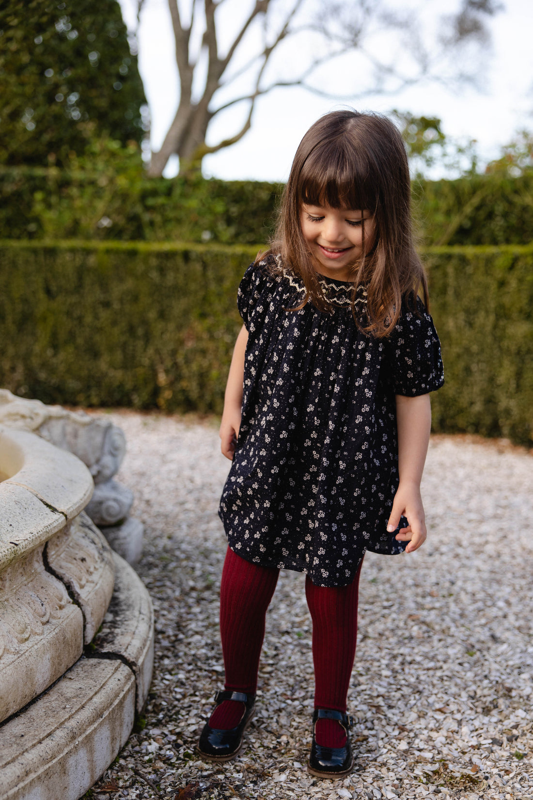 Young girl in a black floral dress standing outdoors on a gravel path with greenery in the background.