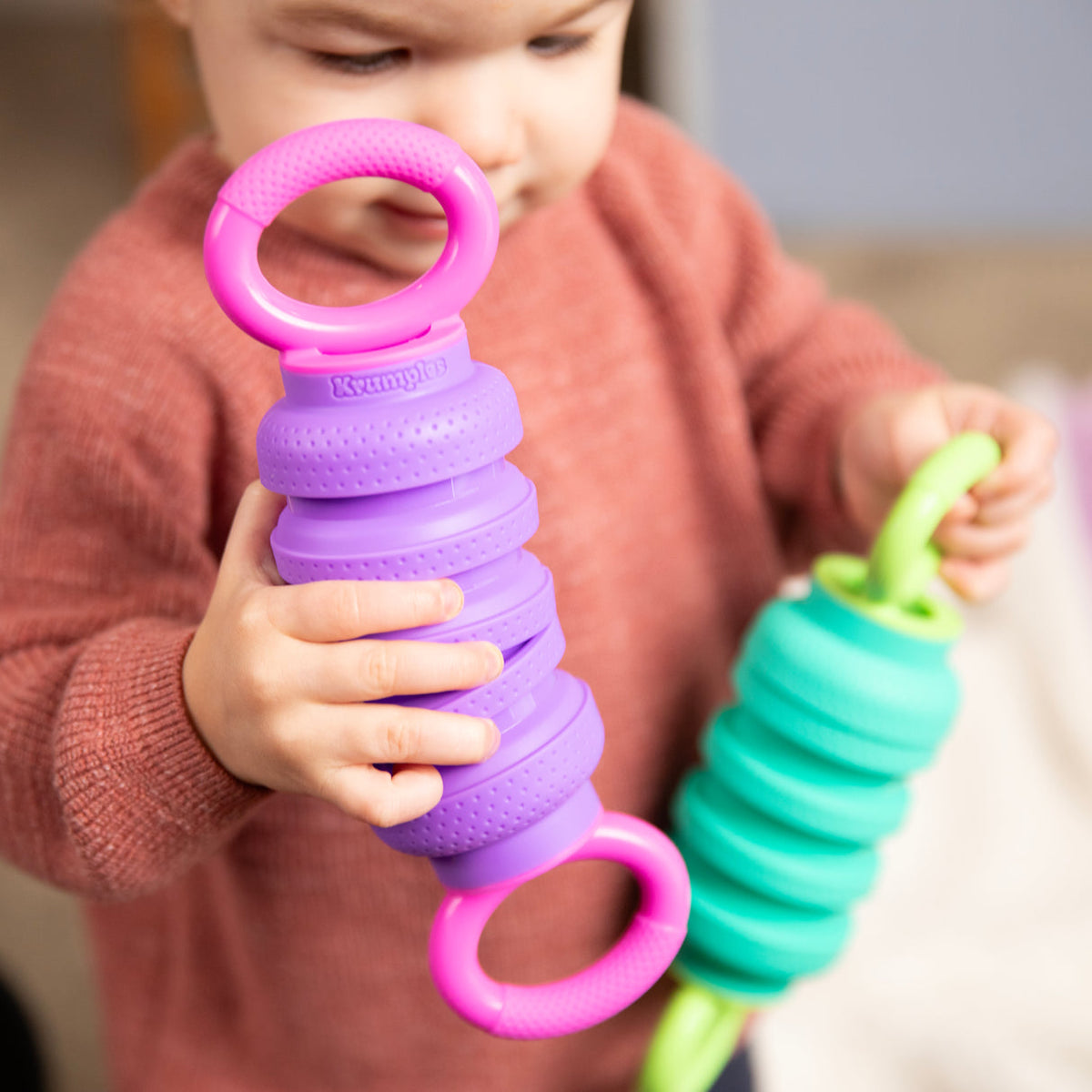 Child holding a purple and pink textured toy with a green and pink toy in the background.
