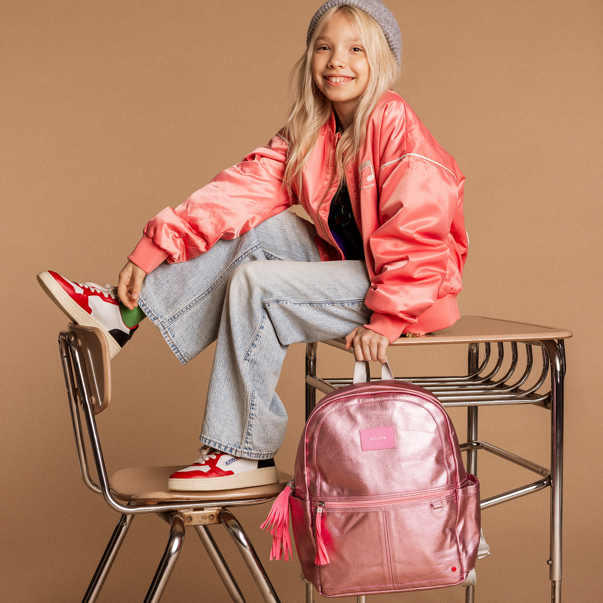 Young girl in pink jacket and jeans sitting on a school desk with a pink backpack, against a brown background