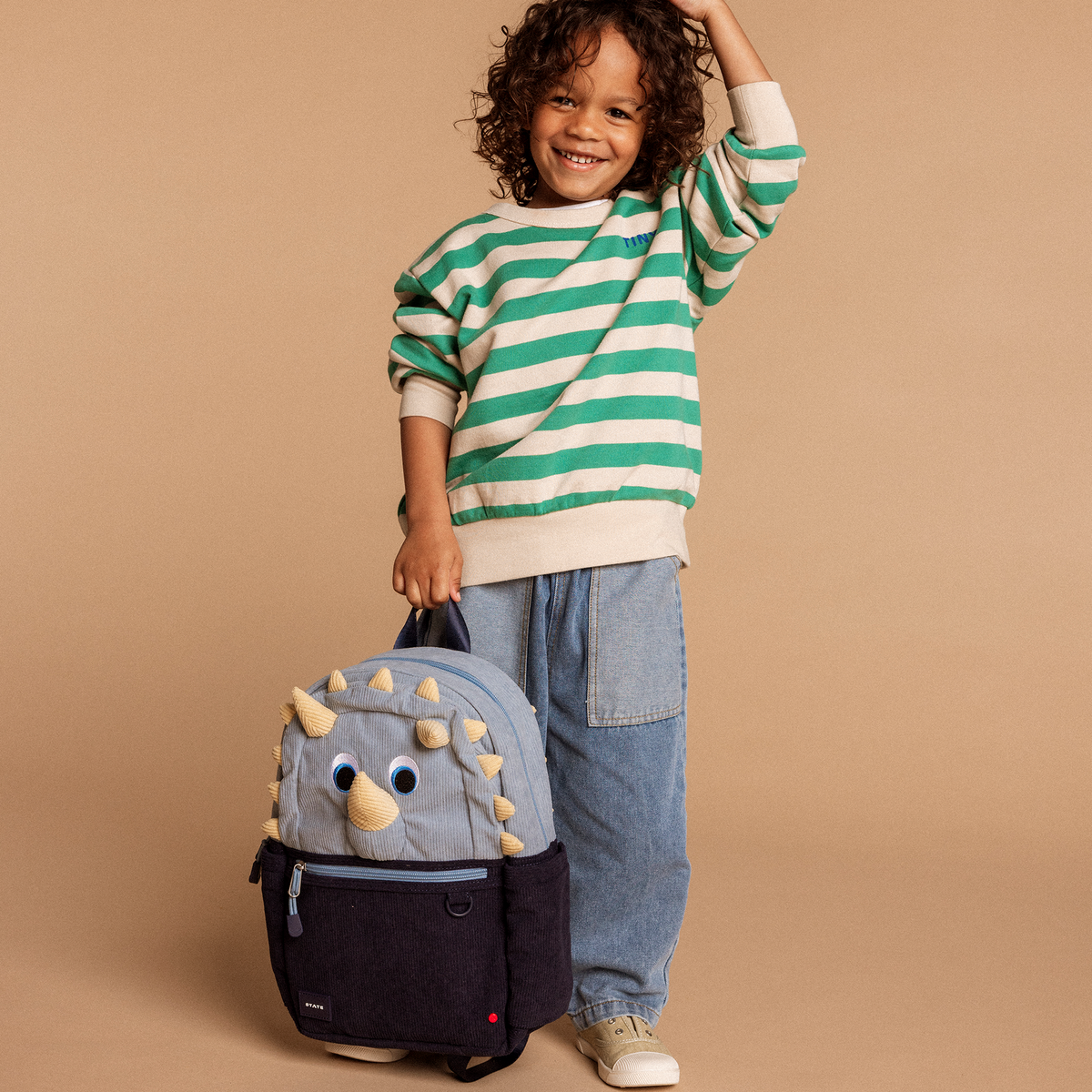 Child wearing a green and white striped sweater with a dinosaur backpack on a beige background