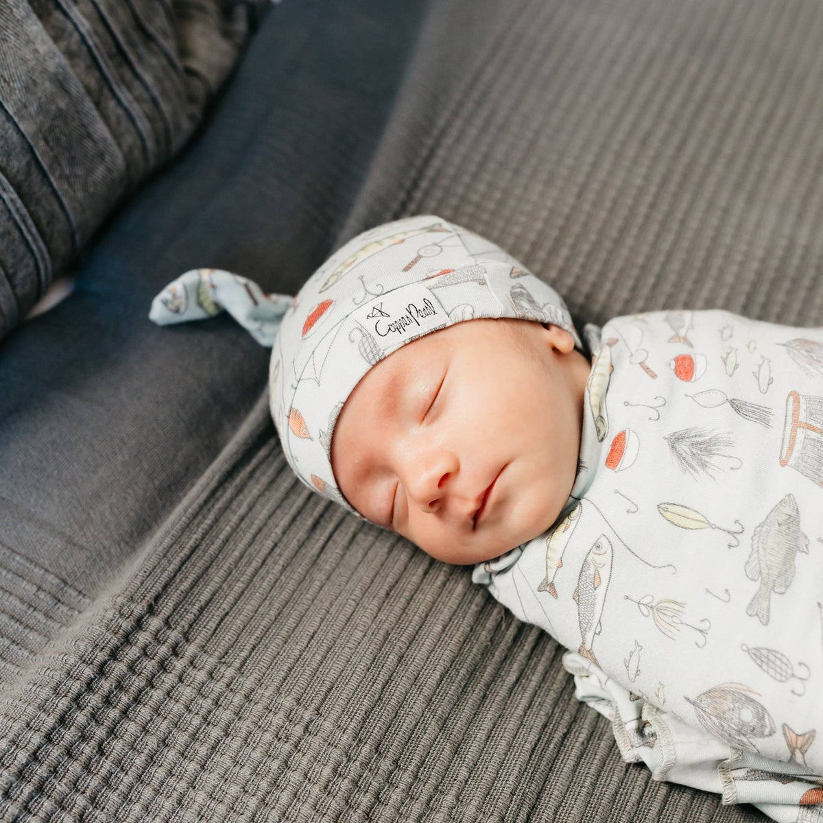 Newborn baby sleeping on a textured surface wearing a patterned hat and swaddle.