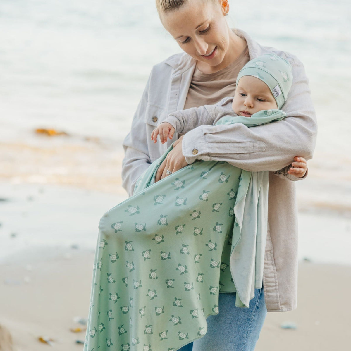 Woman holding a baby wrapped in a green patterned wrap on a beach.