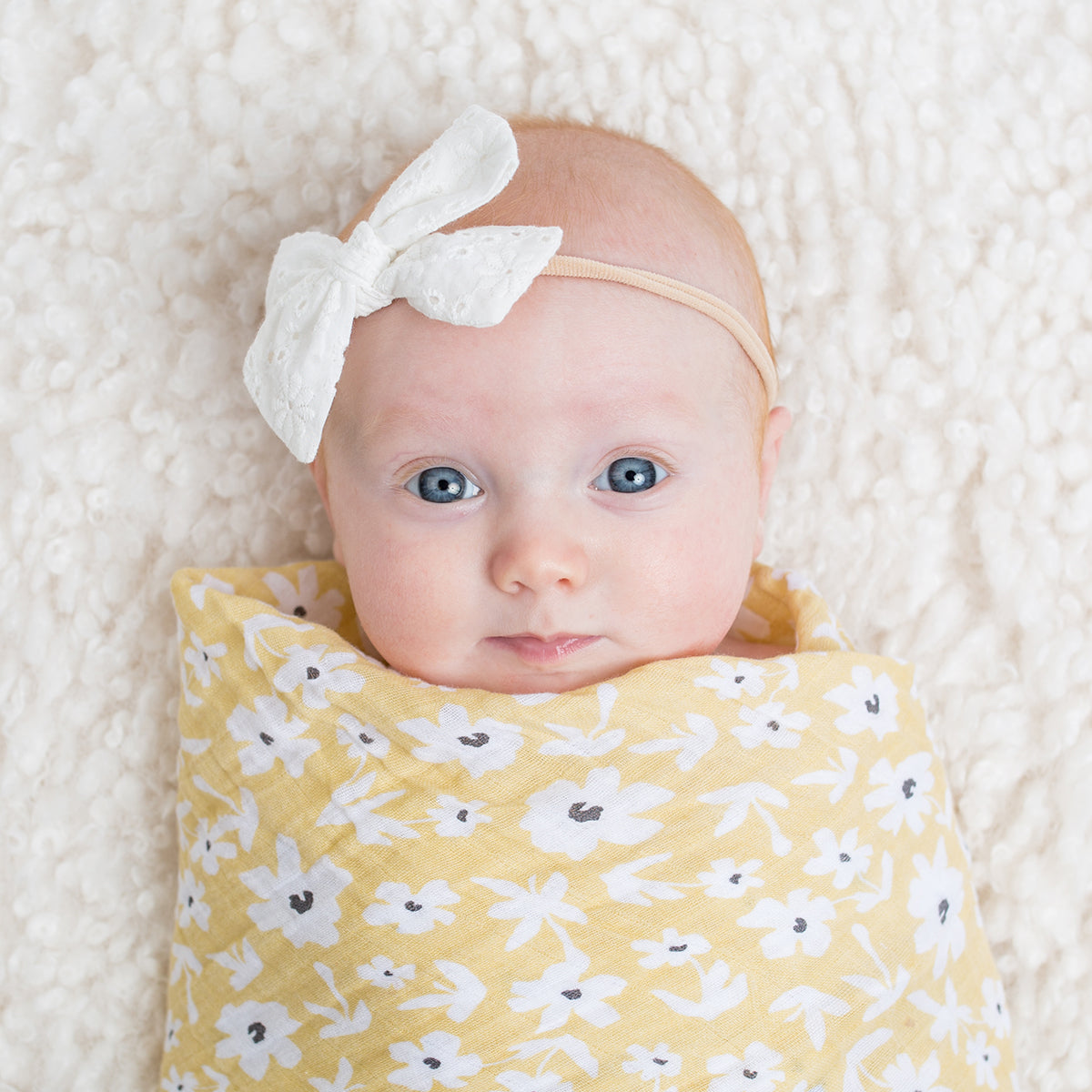 Baby wrapped in a yellow floral blanket with a white bow headband on a soft white background