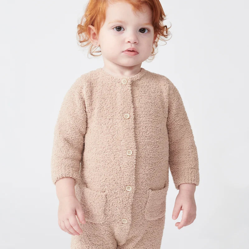 A toddler with reddish-brown hair stands facing forward, wearing a long-sleeve fuzzy romper in a warm pecan brown color by 7AM Enfant, against a plain white background.