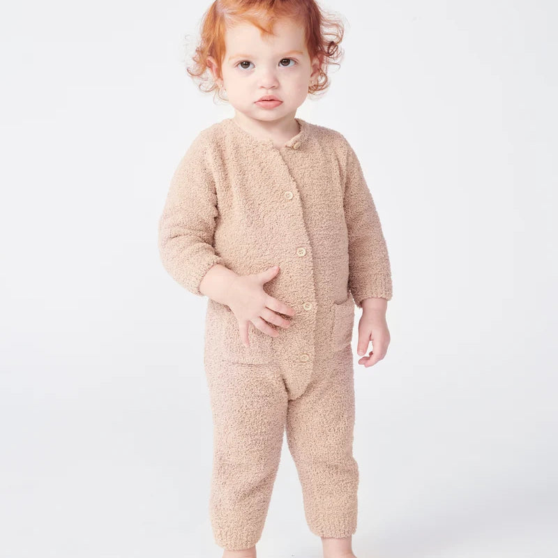 A toddler with reddish-brown hair stands facing forward, wearing a long-sleeve fuzzy romper in a warm pecan brown color by 7AM Enfant, against a plain white background.