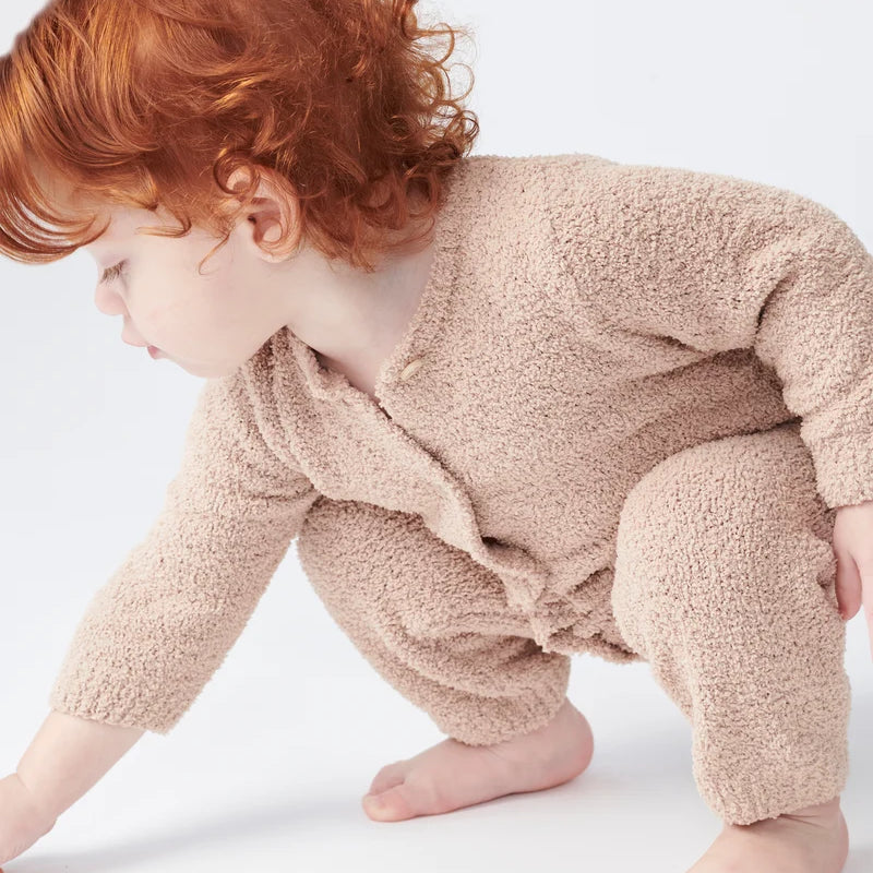 A toddler with reddish-brown hair is crawling on a white surface, wearing a long-sleeve fuzzy romper in a warm pecan brown color by 7AM Enfant.